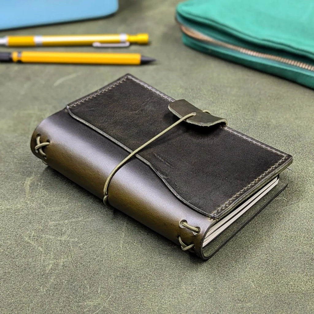 A dark green leather traveler's notebook cover with a live edge front pocket, and an elastic closure strap with a pen loop, placed on a textured green surface with yellow pens and a teal pouch in the background.