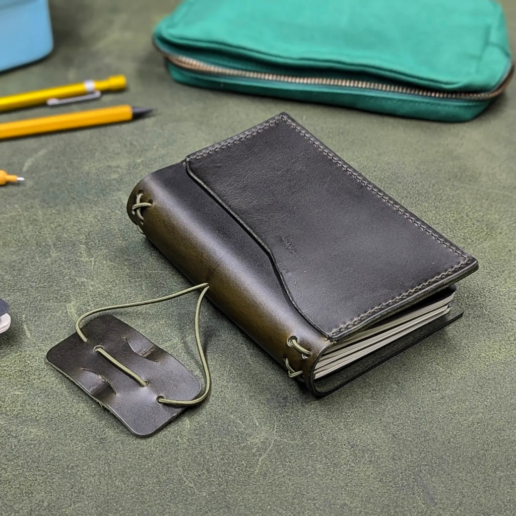 A dark green leather traveler's notebook cover, partially open to show interior notebooks, with a live edge front pocket, and an elastic closure strap with a pen loop, placed on a textured green surface with pencils and a teal pouch in the background.