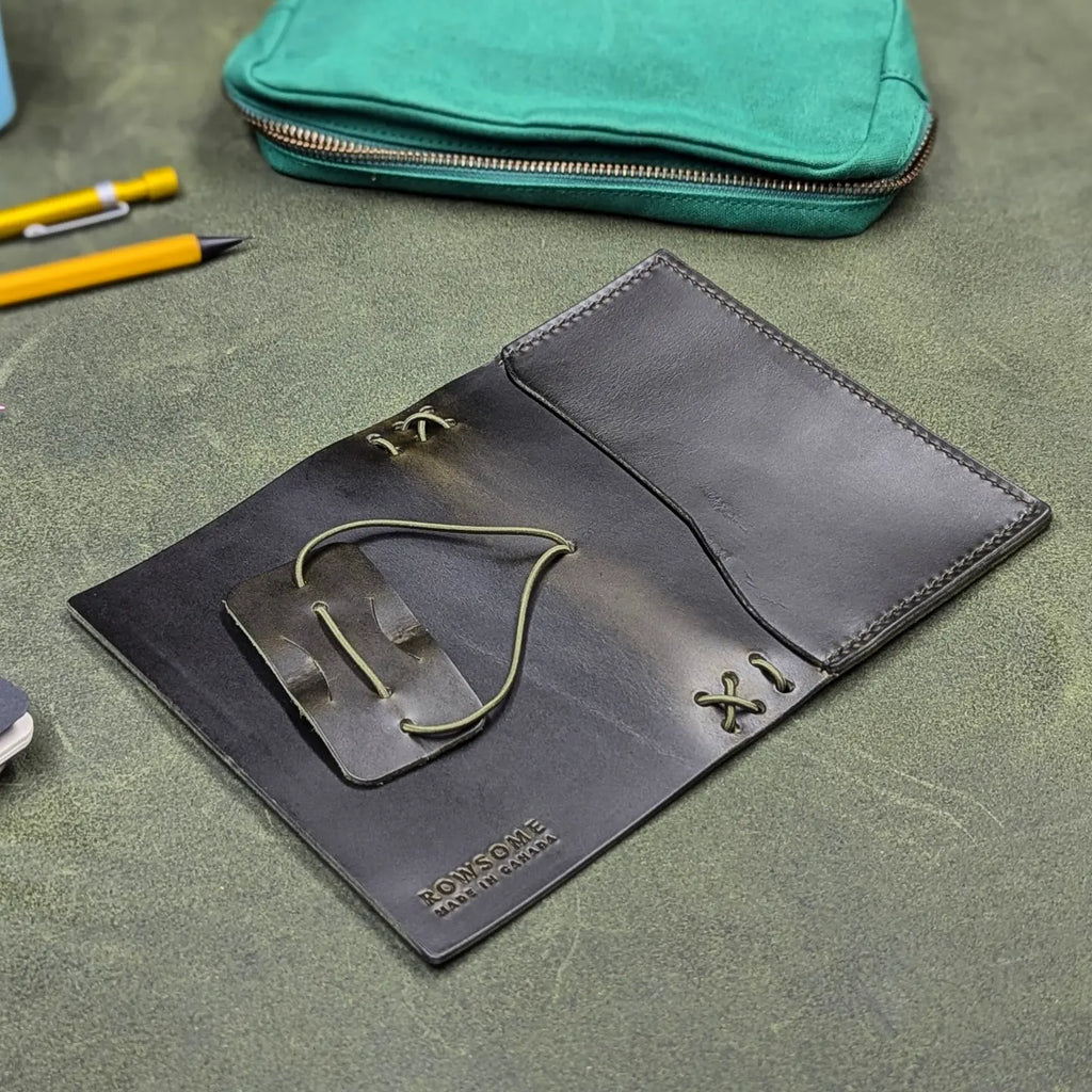 A dark green leather traveler's notebook cover with a live edge front pocket, and an elastic closure strap with a pen loop, shown open, inside-down, on a textured green surface. A teal zip pouch, yellow pen, and partial object are visible in the background as props.