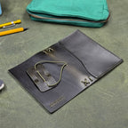 A dark green leather traveler's notebook cover with a live edge front pocket, and an elastic closure strap with a pen loop, shown open, inside-down, on a textured green surface. A teal zip pouch, yellow pen, and partial object are visible in the background as props.