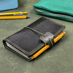 A dark green leather traveler's notebook cover with a live edge front pocket, and an elastic closure strap with a pen loop with a yellow pen inserted. The notebook sits on a textured green surface with other stationery items blurred in the background.