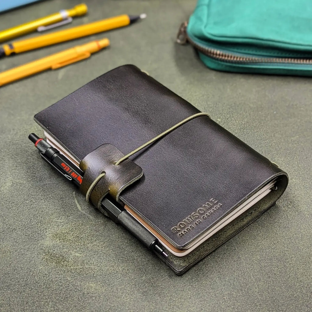 A dark green leather notebook with a wrap-around elastic closure, featuring the debossed brand name 'ROWSOME' on the back cover. A black pen is tucked into the pen loop.