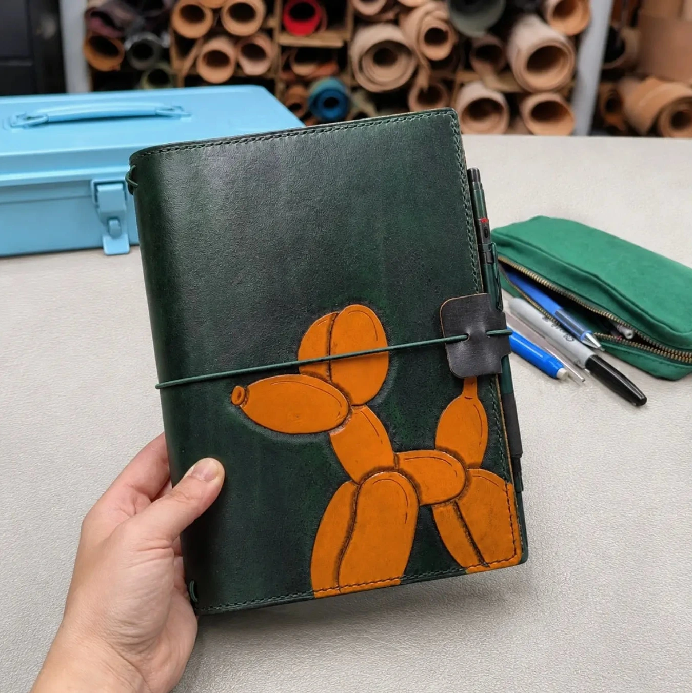 A hand holds a dark green leather journal featuring an orange balloon dog design on the cover, secured with a green elastic band closure, set against a desk background with stationery and a blue case.
