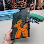 A hand holds a dark green leather notebook cover featuring an orange balloon dog design. The cover has a green elastic closure band and a pen loop on the side.