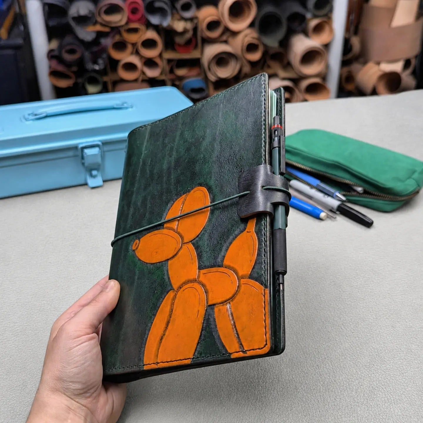 A hand holds a dark green leather notebook cover featuring an orange balloon dog design. The cover has a green elastic closure band and a pen loop on the side.
