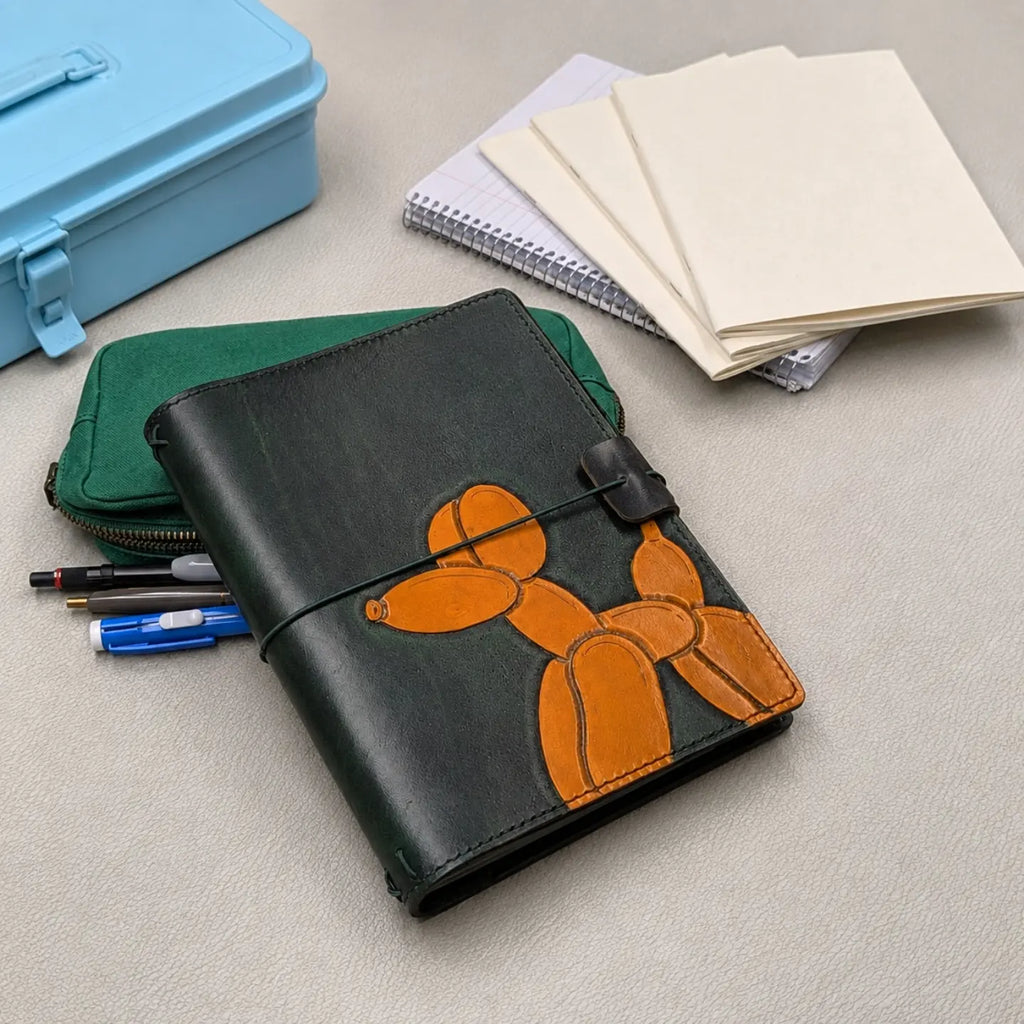 A dark green leather traveler's notebook with an orange balloon dog design, shown on a light grey surface with a green zip pouch, pens, notebooks, and a light blue case nearby.