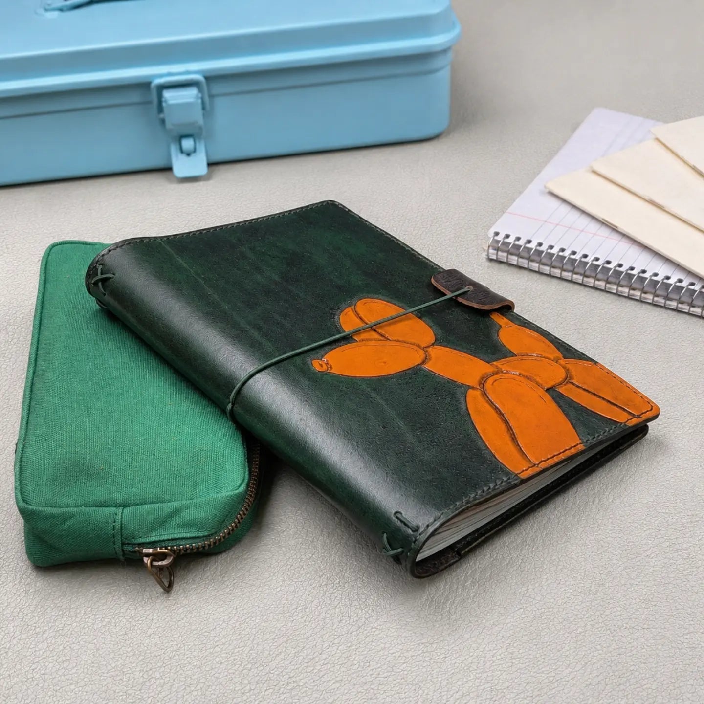 A dark green leather traveler's notebook with an orange balloon dog design, shown on top of a green canvas zip pouch. The scene is styled with a light blue metal case and a spiral notebook in the background.