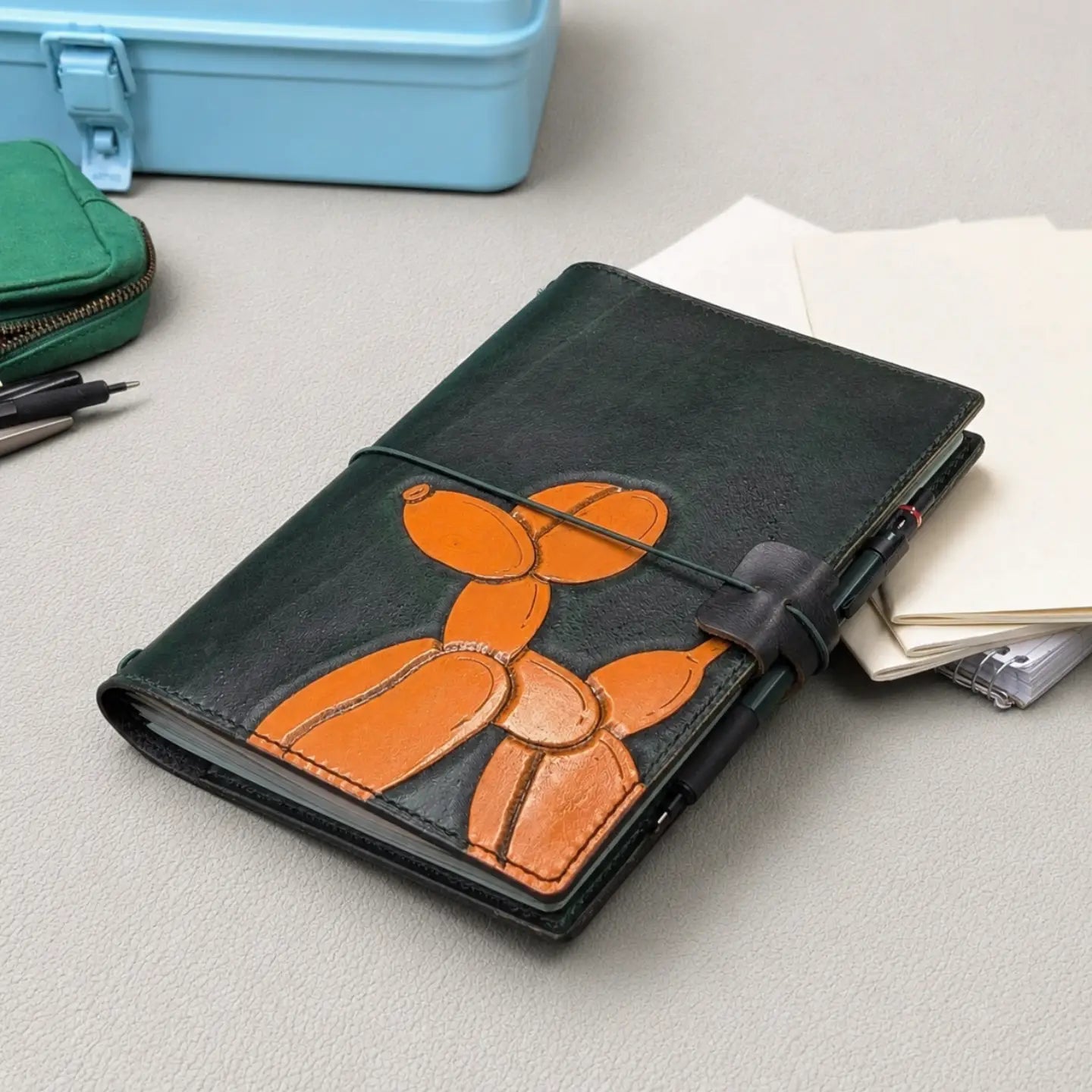 A dark green leather traveler's notebook with an orange balloon dog design, secured with a green elastic closure and a black pen loop, shown on a light gray surface next to papers and stationery.
