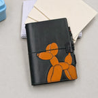 A dark green leather traveler's notebook with an orange balloon dog design on the front, secured with a green closure elastic band. It is styled on a light grey surface with cream-coloured notebooks and a spiral notebook in the background.
