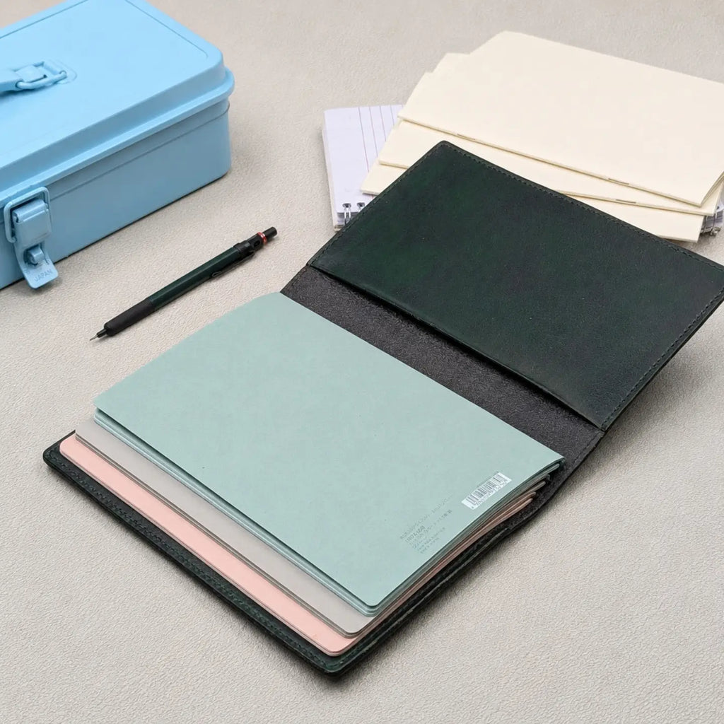 A dark green leather traveler's notebook shown open, revealing multiple pastel-colored notebooks inside, including mint green, pink, and mauve. A light blue metal case, a black pen, and cream notebooks are arranged on a neutral grey textured surface.