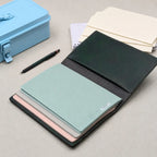 A dark green leather traveler's notebook shown open, revealing multiple pastel-colored notebooks inside, including mint green, pink, and mauve. A light blue metal case, a black pen, and cream notebooks are arranged on a neutral grey textured surface.
