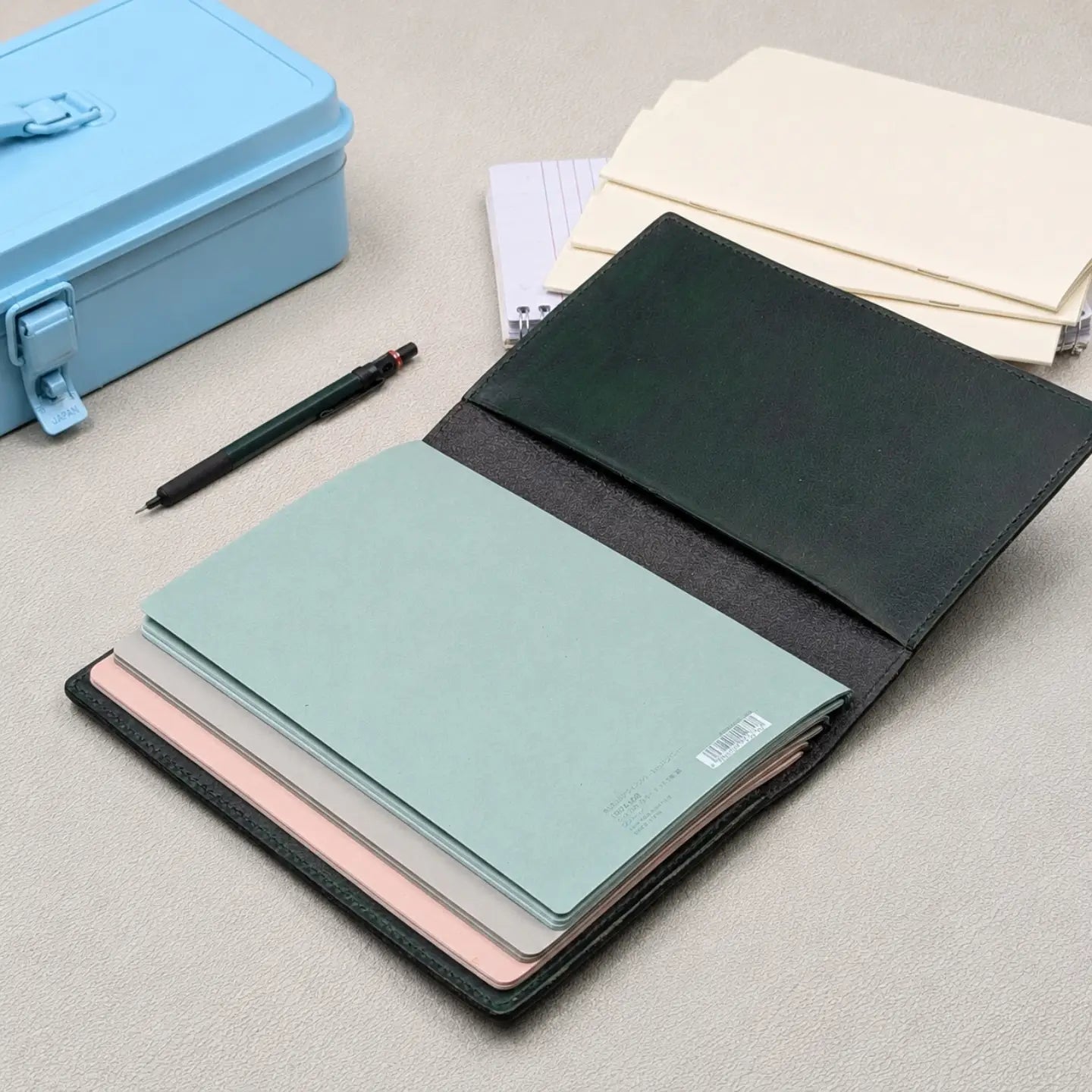 A dark green leather traveler's notebook shown open, revealing multiple pastel-colored notebooks inside, including mint green, pink, and mauve. A light blue metal case, a black pen, and cream notebooks are arranged on a neutral grey textured surface.