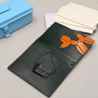 A dark green leather notebook cover featuring an orange balloon dog design on the cover, displayed on a light grey surface with a blue metal case and cream-coloured papers in the background.