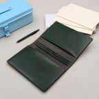 A dark green leather traveler's notebook shown open, showing three sets of elastic bands for holding notebooks, as well as single interior pockets on both the front and back cover. A light blue metal case, a black pen, and cream notebooks are arranged on a neutral grey textured surface.