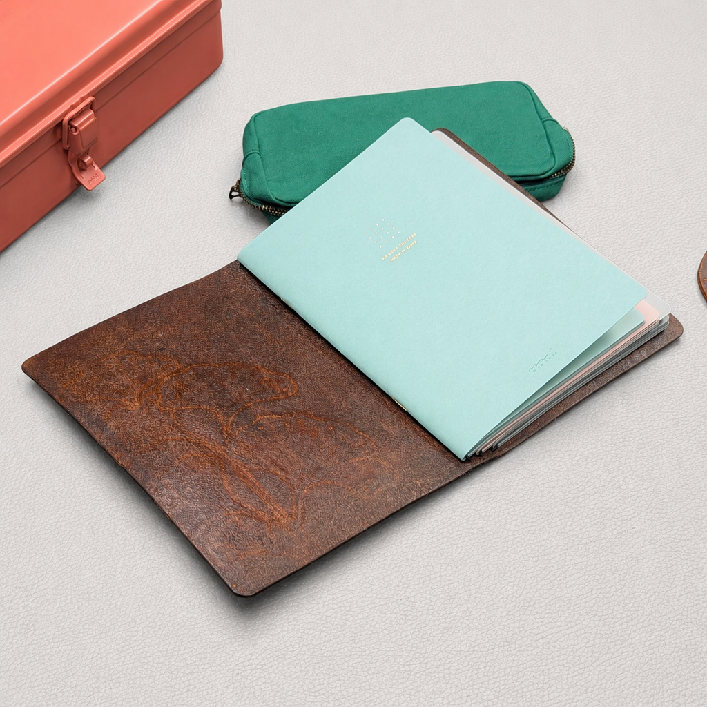 Brown leather notebook cover open holding three notebook inserts with pencil case and toolbox staged around
