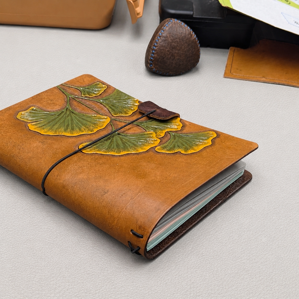 Brown leather notebook cover with green and yellow ginkgo design and a black elastic band with a brown leather pen loop 