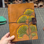 Brown leather notebook cover with green and yellow ginkgo design and a black elastic band with a brown leather pen loop 