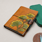 Brown leather notebook cover with green and yellow ginkgo design and a black elastic band with a brown leather pen loop 