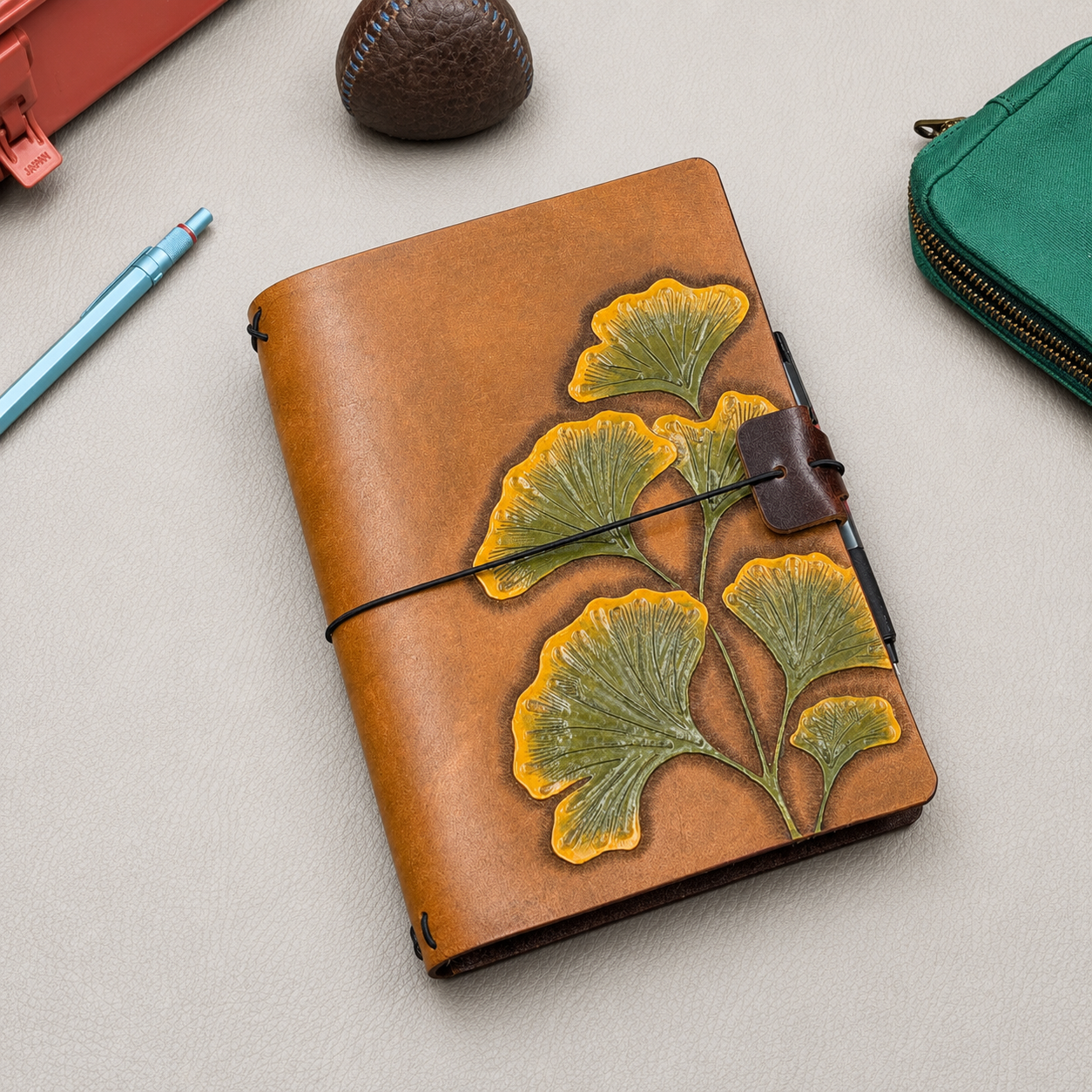 Brown leather notebook cover with green and yellow ginkgo design and a black elastic band with a brown leather pen loop 